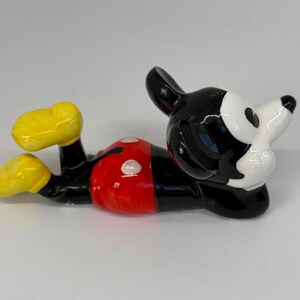 Puede incluir: Figura de cer&aacute;mica de Mickey Mouse tumbado. La figura tiene un cuerpo negro, pantalones cortos rojos con botones blancos, zapatos amarillos y una cara blanca con orejas negras. Un art&iacute;culo de colecci&oacute;n.