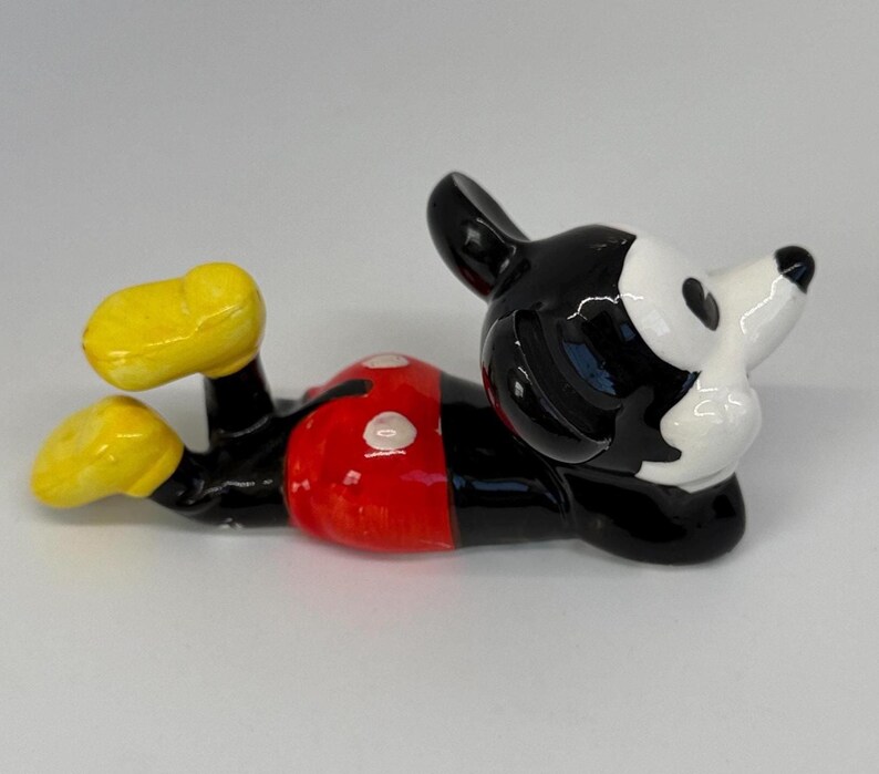 Puede incluir: Figura de cer&aacute;mica de Mickey Mouse tumbado. La figura tiene un cuerpo negro, pantalones cortos rojos con botones blancos, zapatos amarillos y una cara blanca con orejas negras. Un art&iacute;culo de colecci&oacute;n.
