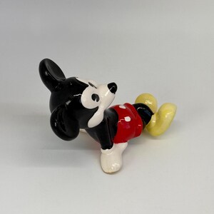 Puede incluir: Una figura de cer&aacute;mica de Mickey Mouse en posici&oacute;n reclinada. La figura presenta orejas y cabeza negras, cara blanca, pantalones cortos rojos con botones blancos, piernas negras y zapatos amarillos. La figura est&aacute; sobre una superficie blanca.