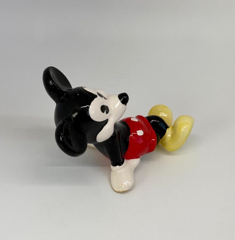 Puede incluir: Una figura de cer&aacute;mica de Mickey Mouse en posici&oacute;n reclinada. La figura presenta orejas y cabeza negras, cara blanca, pantalones cortos rojos con botones blancos, piernas negras y zapatos amarillos. La figura est&aacute; sobre una superficie blanca.
