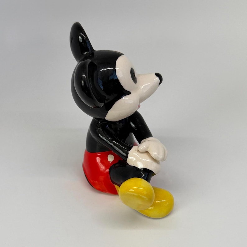 Op de afbeelding: Een keramieken beeldje van Mickey Mouse, met een zwart lichaam, een wit gezicht en een rode broek. Het beeldje heeft gele schoenen en witte handschoenen, in een klassieke zithouding. Een verzamelobject.