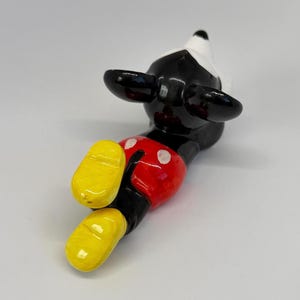 Puede incluir: Una figura de cer&aacute;mica de Mickey Mouse, tumbada boca arriba. La figura tiene un cuerpo negro, una cara blanca y grandes orejas negras. Los pantalones cortos de Mickey son rojos con botones blancos y sus zapatos son amarillos. La figura es brillante.