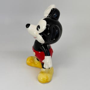 以下が含まれることがあります： ミッキーマウスの陶器製フィギュア。黒い頭と体、白い顔と手袋、赤いズボン、黄色の靴が特徴です。白い帽子をかぶり、立っているポーズです。