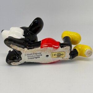 Puede incluir: Una figura de cer&aacute;mica de Mickey Mouse, tumbada boca arriba. La figura tiene cabeza y orejas negras, cara blanca, pantalones cortos rojos y zapatos amarillos. La parte inferior de la figura dice "Walt Disney Productions Copyright".