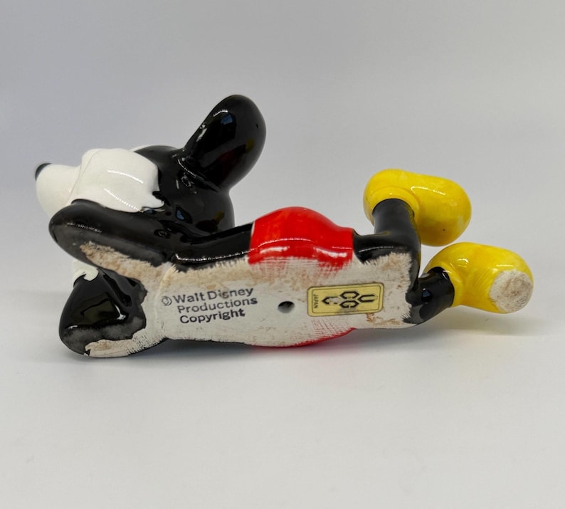 Puede incluir: Una figura de cer&aacute;mica de Mickey Mouse, tumbada boca arriba. La figura tiene cabeza y orejas negras, cara blanca, pantalones cortos rojos y zapatos amarillos. La parte inferior de la figura dice "Walt Disney Productions Copyright".