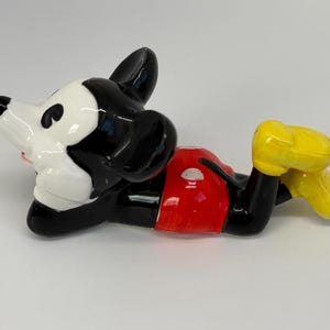 Puede incluir: Una figura de cer&aacute;mica de Mickey Mouse en posici&oacute;n reclinada. La figura presenta un cuerpo negro, una cara blanca, pantalones cortos rojos y zapatos amarillos. La figura es brillante y parece ser un objeto de colecci&oacute;n.