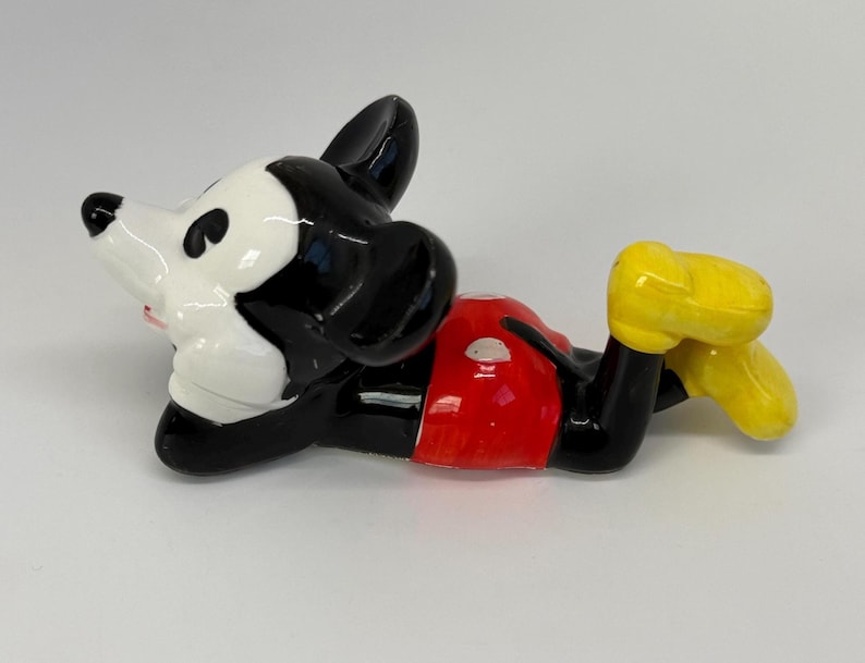 Puede incluir: Una figura de cer&aacute;mica de Mickey Mouse en posici&oacute;n reclinada. La figura presenta un cuerpo negro, una cara blanca, pantalones cortos rojos y zapatos amarillos. La figura es brillante y parece ser un objeto de colecci&oacute;n.