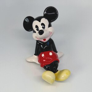 Puede incluir: Una figura de cer&aacute;mica de Mickey Mouse, con orejas negras, una chaqueta negra, pantalones cortos rojos y zapatos amarillos. La figura tiene una cara y manos blancas, con una expresi&oacute;n sonriente. La figura est&aacute; sentada.