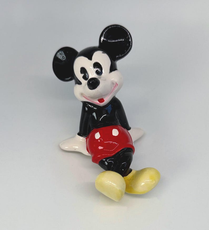 Puede incluir: Una figura de cer&aacute;mica de Mickey Mouse, con orejas negras, una chaqueta negra, pantalones cortos rojos y zapatos amarillos. La figura tiene una cara y manos blancas, con una expresi&oacute;n sonriente. La figura est&aacute; sentada.