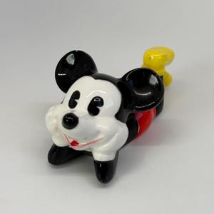 Puede incluir: Una figura de cer&aacute;mica de Mickey Mouse en posici&oacute;n tumbada. La figura tiene orejas negras, cara blanca, pies negros, torso rojo y cola amarilla. La figura es un objeto de colecci&oacute;n.