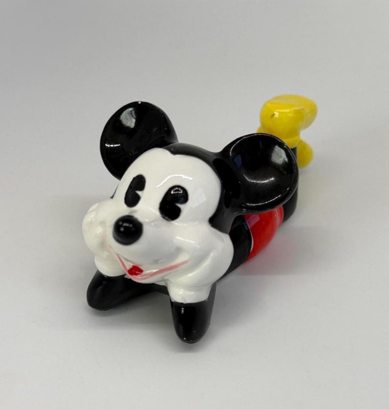 Puede incluir: Una figura de cer&aacute;mica de Mickey Mouse en posici&oacute;n tumbada. La figura tiene orejas negras, cara blanca, pies negros, torso rojo y cola amarilla. La figura es un objeto de colecci&oacute;n.