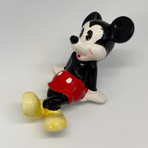 Puede incluir: Figura de cer&aacute;mica de Mickey Mouse en una pose juguetona. La figura es predominantemente negra, con pantalones cortos rojos y guantes blancos. Las orejas son grandes y negras, y los zapatos son amarillos. La figura tiene un acabado brillante sobre un fondo blanco.