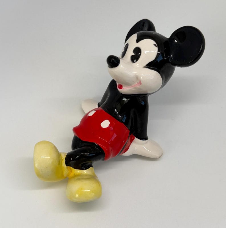 Puede incluir: Figura de cer&aacute;mica de Mickey Mouse en una pose juguetona. La figura es predominantemente negra, con pantalones cortos rojos y guantes blancos. Las orejas son grandes y negras, y los zapatos son amarillos. La figura tiene un acabado brillante sobre un fondo blanco.