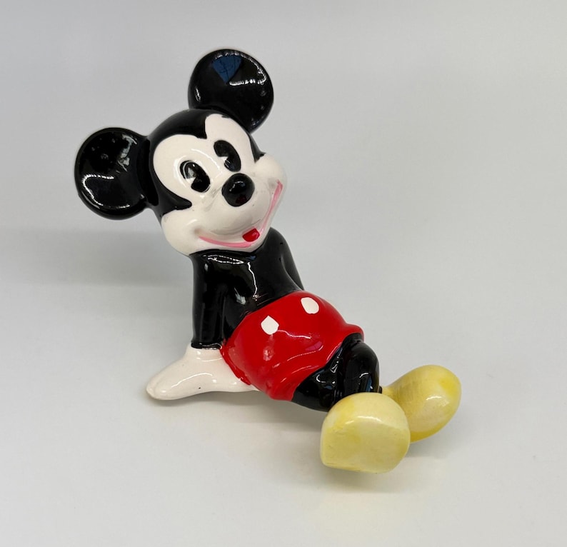 Puede incluir: Figura de cer&aacute;mica de Mickey Mouse, con cuerpo negro, cara blanca y pantalones cortos rojos. La figura tiene zapatos amarillos y est&aacute; en posici&oacute;n sentada. Objeto de colecci&oacute;n.