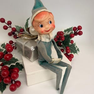 May include: A vintage-style elf figurine with a teal felt suit and matching pointed hat, featuring a painted face with rosy cheeks and blue eyes. The elf sits beside a small silver gift box, surrounded by artificial holly branches with red berries.