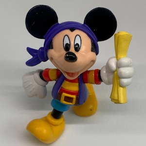 May include: A Mickey Mouse figurine dressed as a pirate, wearing a purple bandana, vest, and yellow boots. The figure is holding a yellow scroll and has white gloves. The figure has black ears and a smiling expression.