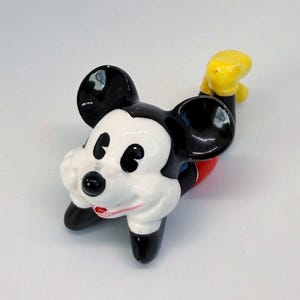 Puede incluir: Figura de cer&aacute;mica de Mickey Mouse en posici&oacute;n reclinada. La figura presenta orejas negras, nariz negra y cara blanca. Mickey viste pantalones cortos rojos y guantes amarillos. La figura est&aacute; sobre una superficie blanca.