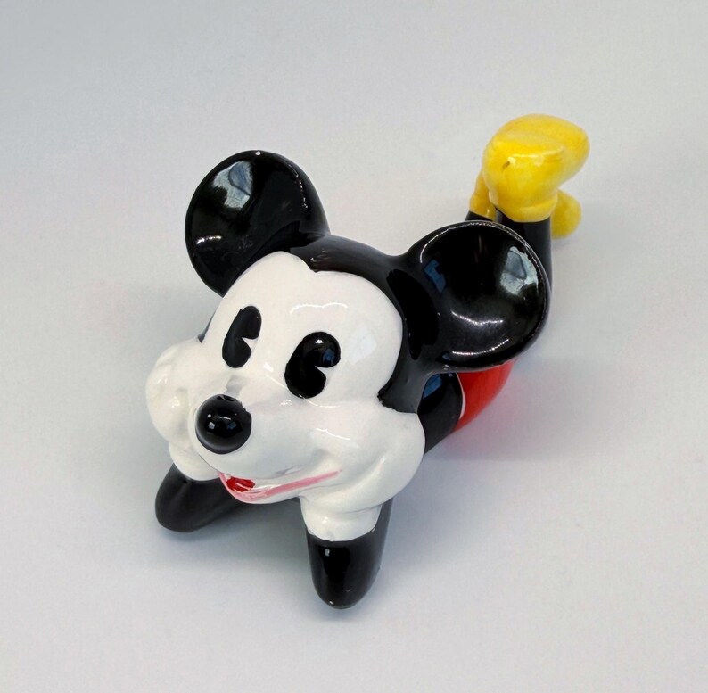 Puede incluir: Figura de cer&aacute;mica de Mickey Mouse en posici&oacute;n reclinada. La figura presenta orejas negras, nariz negra y cara blanca. Mickey viste pantalones cortos rojos y guantes amarillos. La figura est&aacute; sobre una superficie blanca.