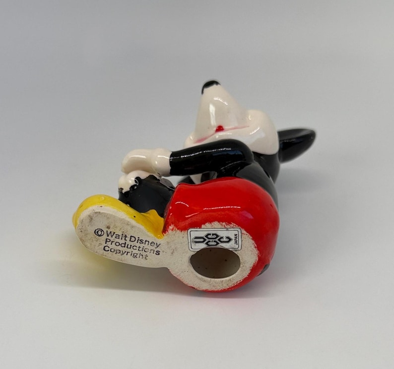 Op de afbeelding: Een keramieken Mickey Mouse beeldje in een liggende houding. De figuur heeft een witte kop, een zwart lichaam, een rode broek en gele schoenen. De basis van het beeldje heeft de tekst "Walt Disney Productions Copyright".