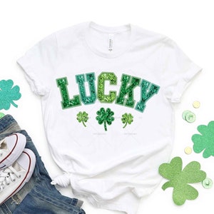 May include: White t-shirt with the word "LUCKY" in green glitter letters. There are three green glitter shamrocks below the word.