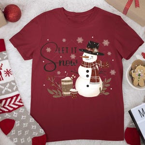 May include: Red t-shirt with a snowman graphic and the text "Let it snow". The snowman is wearing a red and black plaid scarf and a black hat.