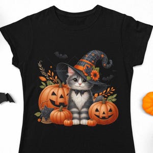 K&ouml;nnte beinhalten: Ein schwarzes T-Shirt mit einer s&uuml;&szlig;en grau-wei&szlig;en Katze, die einen Hexenhut tr&auml;gt und von K&uuml;rbissen umgeben ist. Die Katze sitzt auf einem Stapel K&uuml;rbisse, von denen einer ein Jack-o'-Lantern-Gesicht hat. Der Hintergrund zeigt eine dunkle Waldszene mit herumfliegenden Flederm&auml;usen.