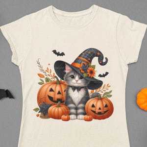 K&ouml;nnte beinhalten: Ein wei&szlig;es T-Shirt mit einer s&uuml;&szlig;en grauen Katze, die einen Hexenhut tr&auml;gt und einen K&uuml;rbis h&auml;lt. Die Katze ist von K&uuml;rbissen und Herbstbl&auml;ttern umgeben.