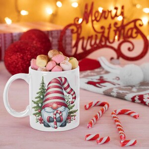 May include: A white ceramic mug filled with marshmallows, featuring a Christmas gnome design with a red and white striped hat and a candy cane. The mug is surrounded by candy canes and festive decorations.