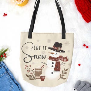 May include: A beige tote bag with black straps features a snowman wearing a red and black plaid scarf and a black hat with a red flower. The bag has the text "Let it Snow" in black script.