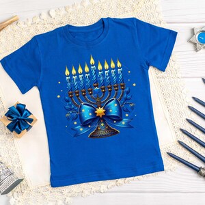 Op de afbeelding: Een blauw T-shirt met een afbeelding van een menorah met negen brandende kaarsen, een blauwe strik en een gouden ster. De menorah is omringd door een bloemmotief.
