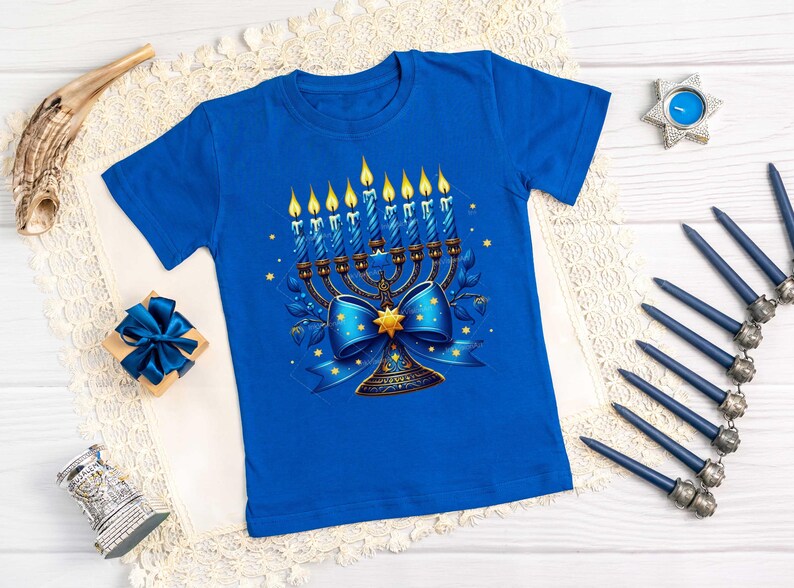 Op de afbeelding: Een blauw T-shirt met een afbeelding van een menorah met negen brandende kaarsen, een blauwe strik en een gouden ster. De menorah is omringd door een bloemmotief.