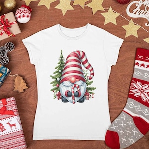 May include: White t-shirt featuring a Christmas gnome with a red and white striped hat, holding a candy cane. The gnome is surrounded by a Christmas tree and festive decorations. The shirt is displayed on a wooden surface with Christmas ornaments.