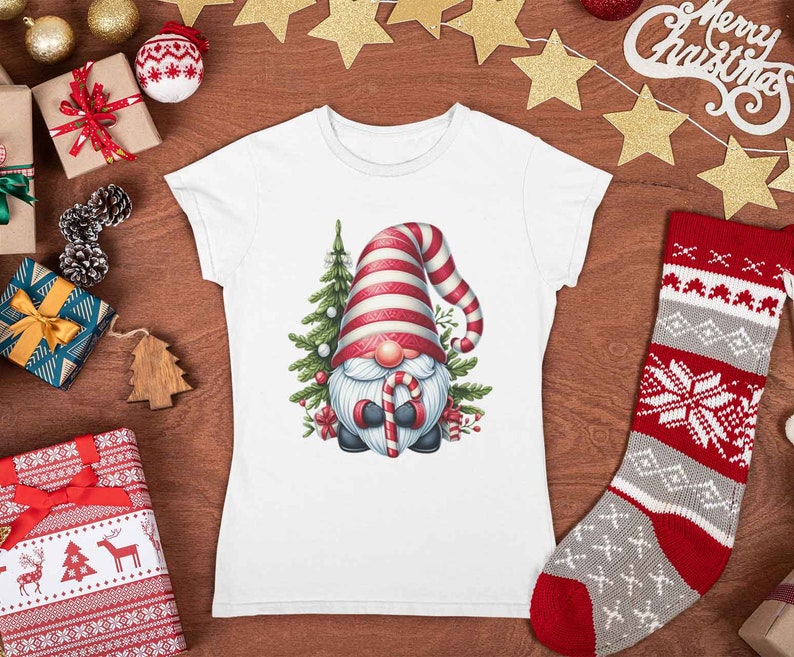 May include: White t-shirt featuring a Christmas gnome with a red and white striped hat, holding a candy cane. The gnome is surrounded by a Christmas tree and festive decorations. The shirt is displayed on a wooden surface with Christmas ornaments.