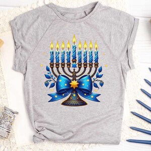 Op de afbeelding: Een grijs T-shirt met een blauwe en gouden menorah met een blauwe strik en sterren. De menorah heeft negen kaarsen, waarvan er zeven branden.