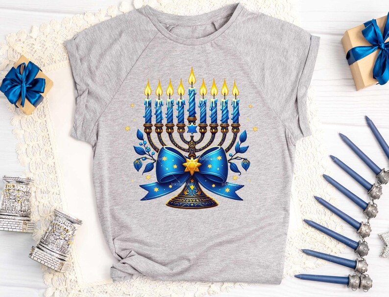 Op de afbeelding: Een grijs T-shirt met een blauwe en gouden menorah met een blauwe strik en sterren. De menorah heeft negen kaarsen, waarvan er zeven branden.