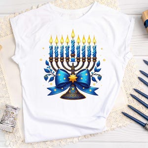 Op de afbeelding: Wit T-shirt met een blauwe en gouden menorah met brandende kaarsen en een blauwe strik. De menorah is versierd met sterren en de strik heeft een gouden ster in het midden.