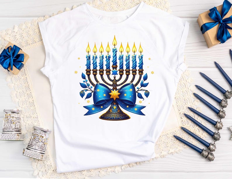 Op de afbeelding: Wit T-shirt met een blauwe en gouden menorah met brandende kaarsen en een blauwe strik. De menorah is versierd met sterren en de strik heeft een gouden ster in het midden.