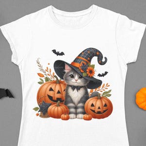 K&ouml;nnte beinhalten: Ein wei&szlig;es T-Shirt mit einer s&uuml;&szlig;en grau-wei&szlig;en Katze, die einen Hexenhut tr&auml;gt und einen K&uuml;rbis h&auml;lt. Die Katze ist von K&uuml;rbissen und Flederm&auml;usen umgeben.