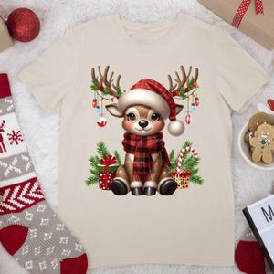 Pode incluir: Uma camiseta bege com uma rena de desenho animado usando uma echarpe xadrez vermelha e preta e um chap&eacute;u de Papai Noel. A rena est&aacute; cercada por luzes de Natal, presentes e bengalas de doces.