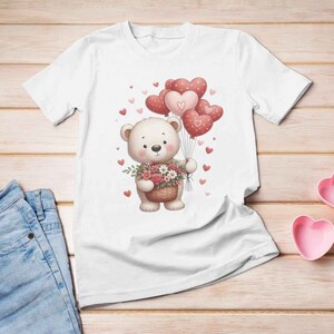 May include: White t-shirt with a cute cartoon bear holding a basket of flowers. The bear is surrounded by pink heart-shaped balloons.