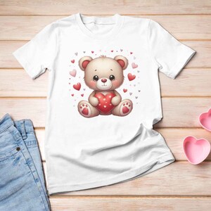 May include: White t-shirt with a cartoon illustration of a brown teddy bear holding a red heart. The bear is surrounded by small pink and red hearts.