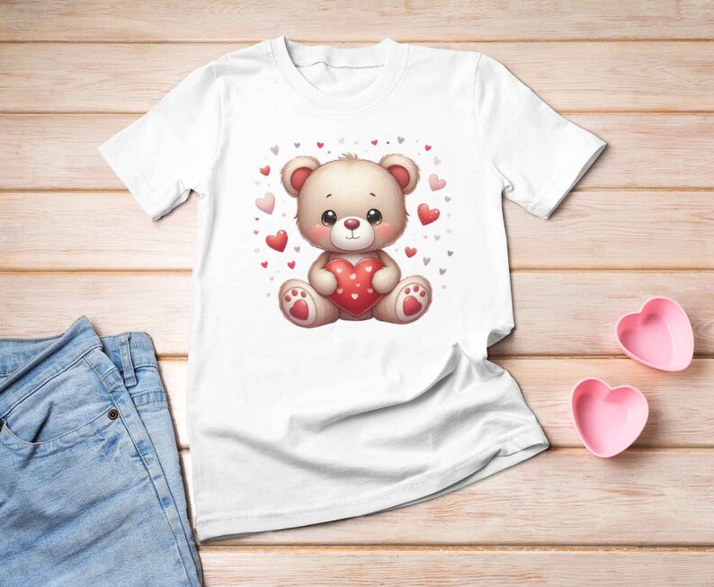 May include: White t-shirt with a cartoon illustration of a brown teddy bear holding a red heart. The bear is surrounded by small pink and red hearts.