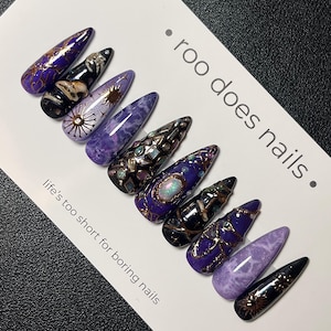 Könnte beinhalten: Ein Set aus zehn langen, mandelförmigen Kunstnägeln. Die Nägel weisen eine Vielzahl von Designs in Lila-, Schwarz- und Goldtönen auf. Einige Nägel haben komplizierte Muster, während andere einen Marmoreffekt aufweisen. Der Text "roo does nails" und "life's too short for boring nails" ist ebenfalls sichtbar.