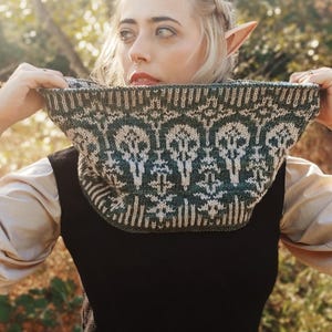 Puede incluir: Un cuello tejido en verde y crema con un patrón repetido de calaveras. El cuello se sostiene, revelando el diseño detallado y la textura del tejido. El fondo está borroso, lo que sugiere un entorno al aire libre. Un accesorio de moda.