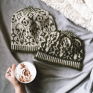 Puede incluir: Dos gorros de punto con un patrón geométrico en verde oliva y crema. Los sombreros se muestran sobre una superficie gris con una manta blanca y esponjosa en el fondo. Una mano sostiene una taza de café con un remolino de crema.