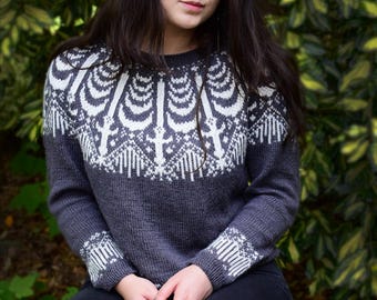 Könnte beinhalten: Ein dunkelgrauer Pullover mit einem weißen geometrischen Muster auf der Brust und an den Manschetten. Der langärmlige Pullover scheint aus einem weichen Strickmaterial zu sein. Die Person trägt eine schwarze Jeans.