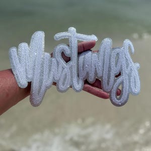 Op de afbeelding: Een zilveren pailletten "Mustangs" woordkunstwerk, vastgehouden in een hand tegen een vage strandachtergrond. De cursieve letters zijn omlijnd met een dikke, getextureerde rand, waardoor een 3D-effect ontstaat. De pailletten vangen het licht.
