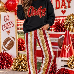 May include: A black long-sleeve sweatshirt with the word Chiefs in red glittery script lettering, paired with straight-leg pants featuring vertical sequined stripes in red, gold, and silver, and white sneakers with a red star detail.
