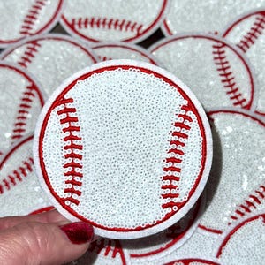 Può includere: Primo piano di un mucchio di toppe a forma di palla da baseball bianche e rosse con paillettes. Le toppe hanno un diametro di circa 7,5 cm e hanno cuciture rosse attorno alle cuciture.