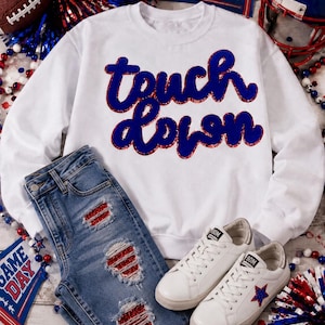 May include: A white crewneck sweatshirt with the text "touch down" in blue glitter script, paired with blue denim jeans featuring distressed patches backed with red glitter fabric, and white sneakers with a blue and red glitter star detail.
